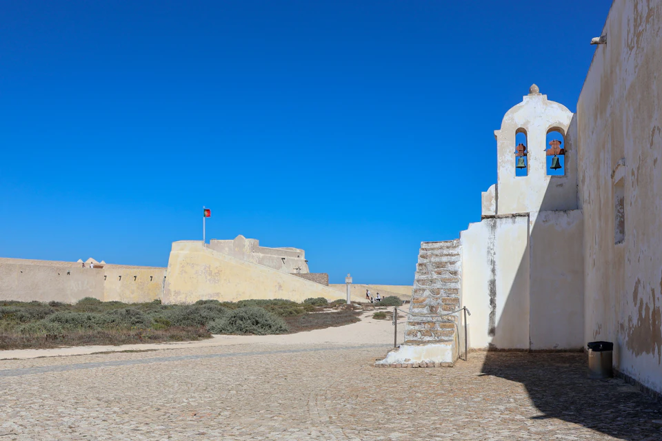 A small white chapel with a bell tower beside a stone fortress under a clear blue sky.