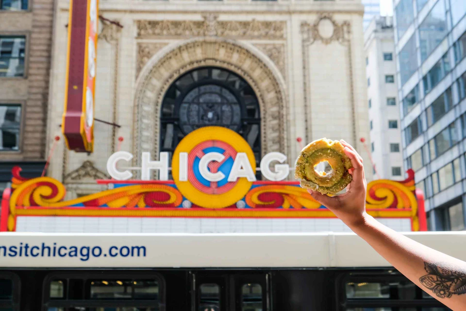A hand holds a donut in front of the iconic Chicago Theater marquee displaying "CHICAGO," with a bus partially visible below.