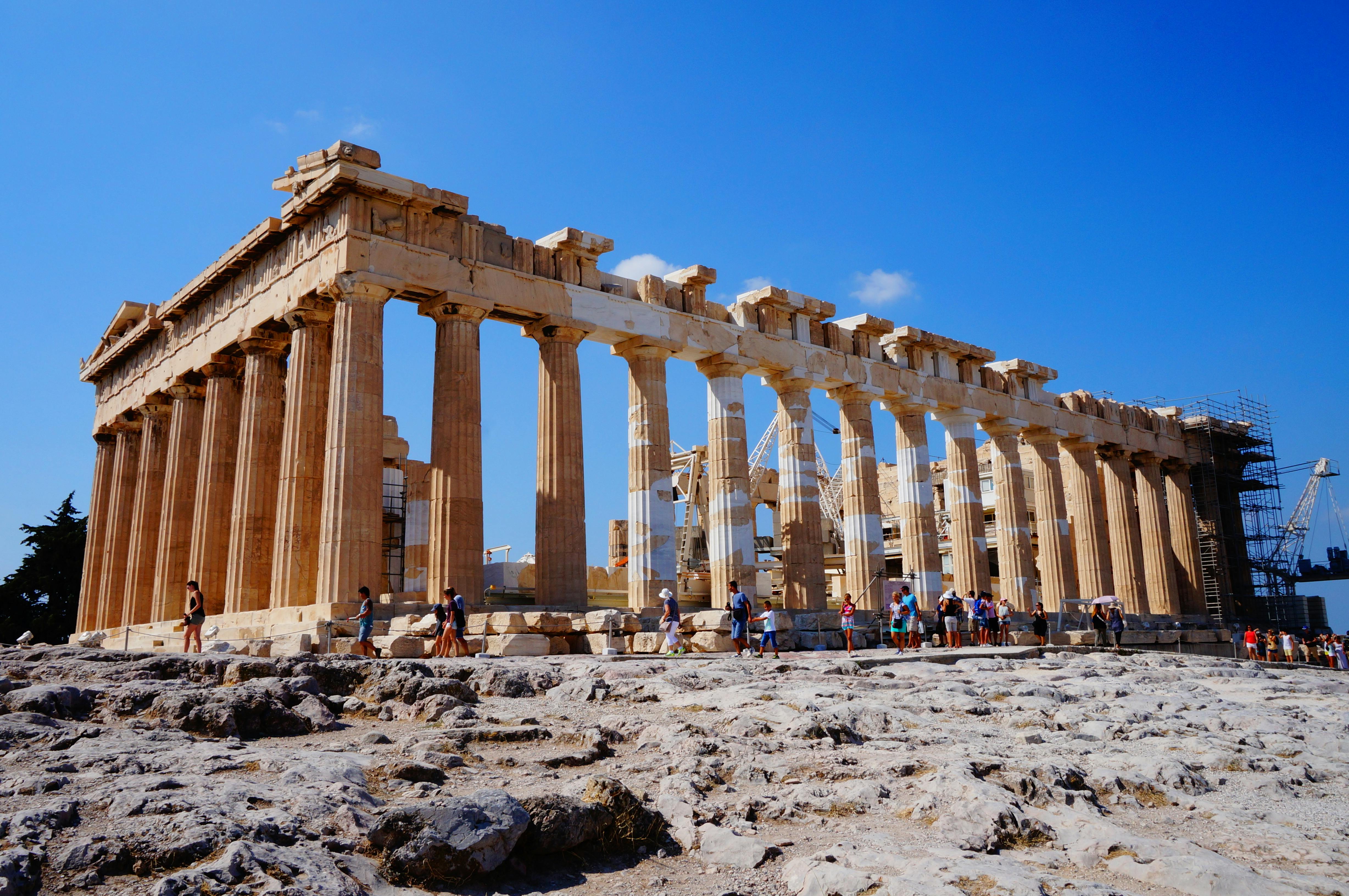 Tourists explore the ancient Parthenon, a large columned temple, under a bright blue sky with scattered clouds.