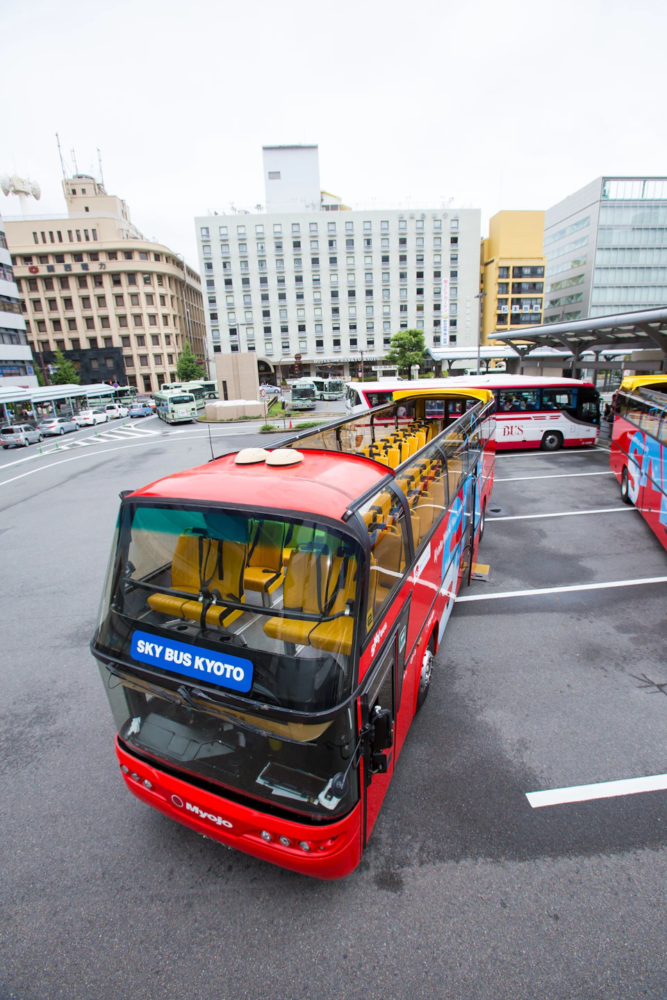 Red, double-decker bus labeled "Sky Bus Kyoto" parked in a bus lot with multiple similar buses and buildings in the background.