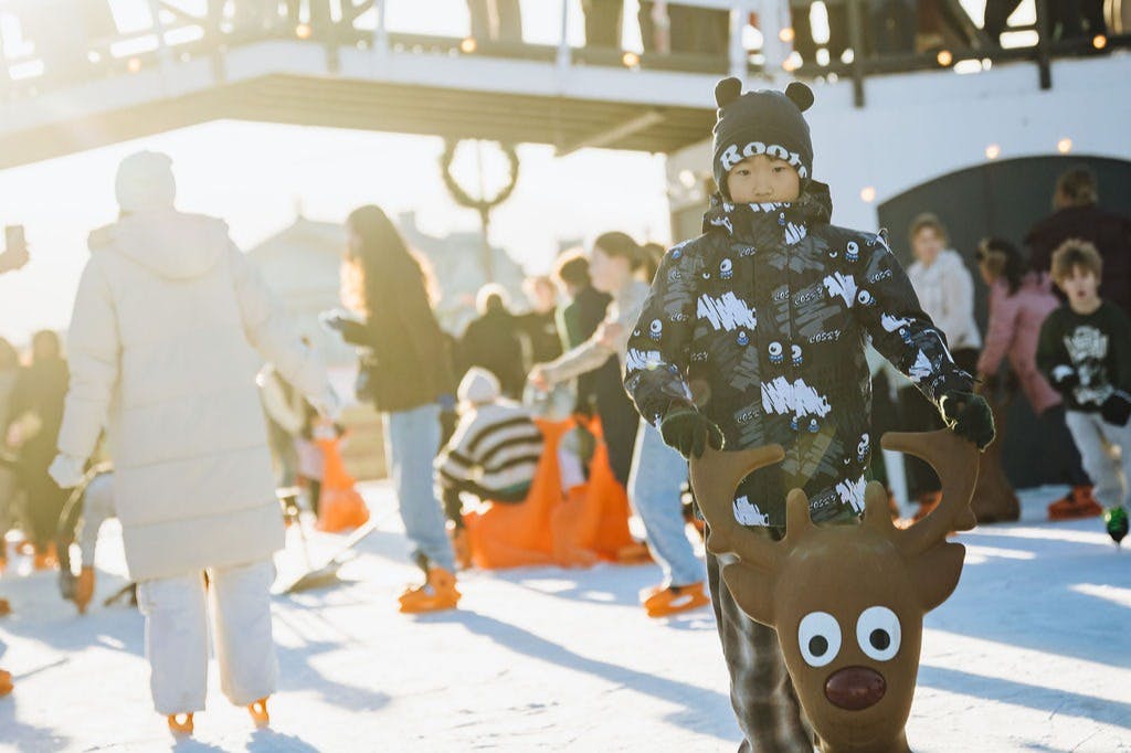 A child in winter clothing skates with a reindeer-shaped aid on an ice rink, surrounded by other skaters.