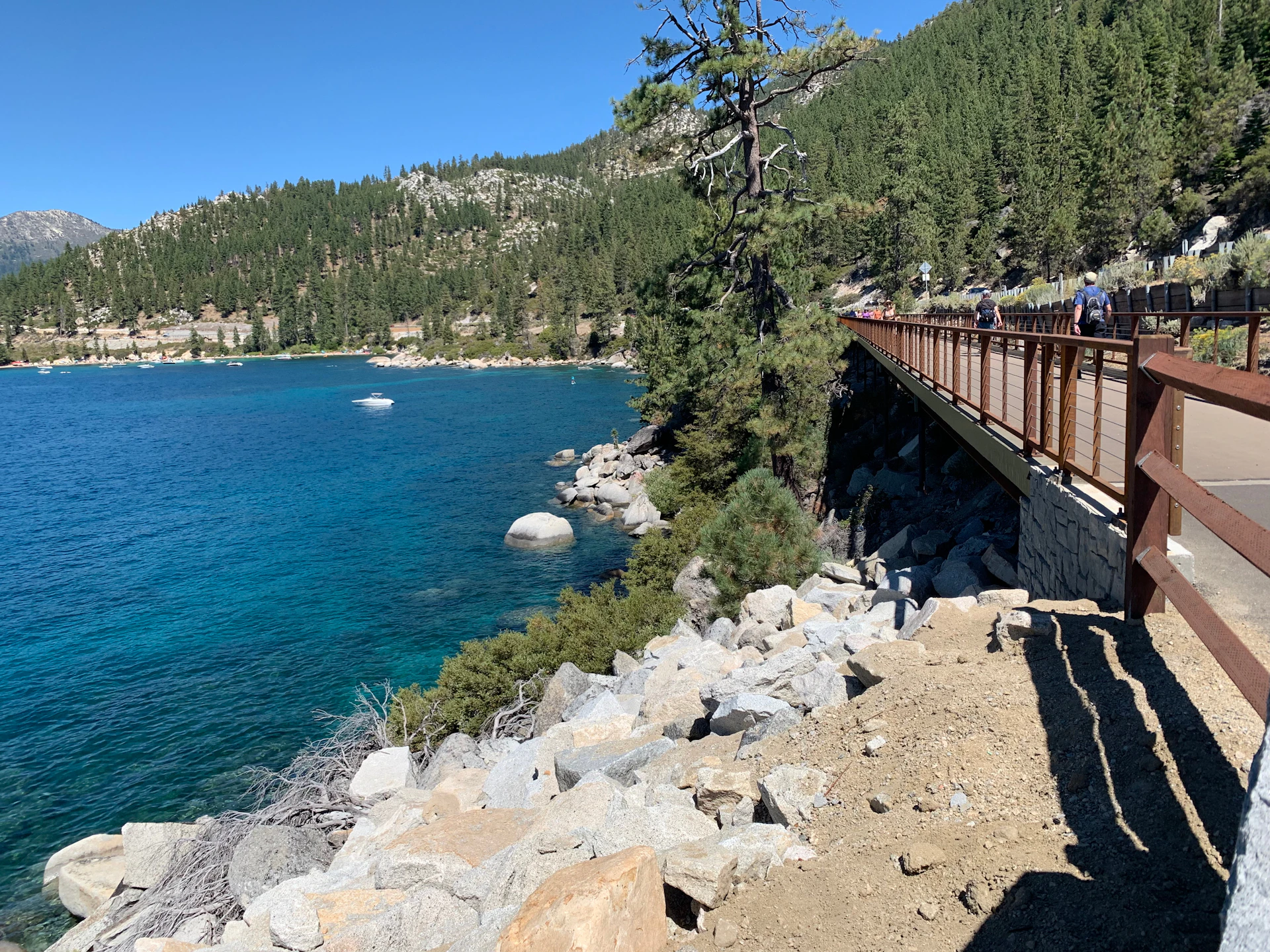 A wooden bridge with people walking on it overlooks a rocky shoreline and a blue lake surrounded by pine-covered hills.