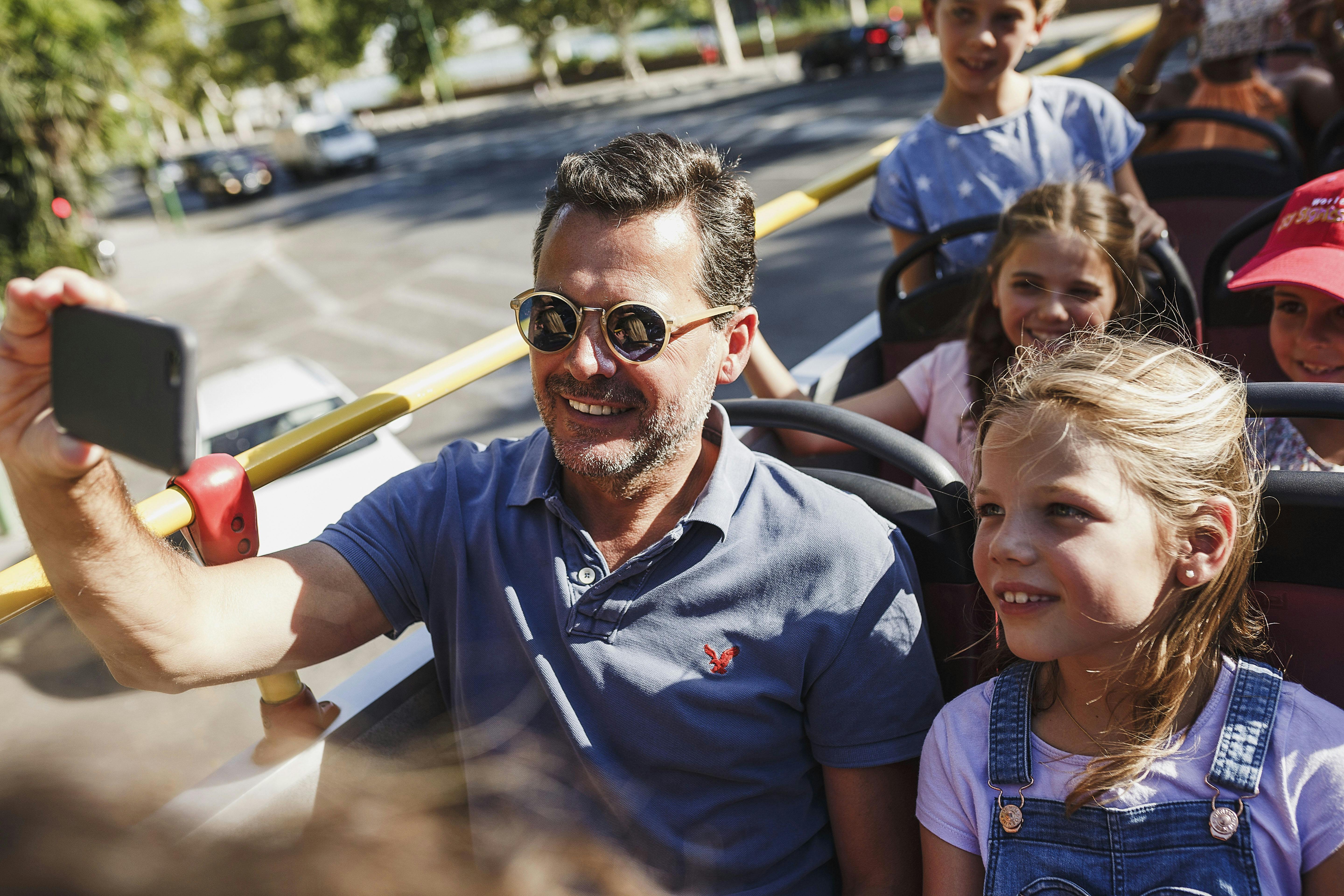 Skupina lidí, včetně dětí, se usmívá a užívá si slunečný den při jízdě autobusem s otevřenou střechou.