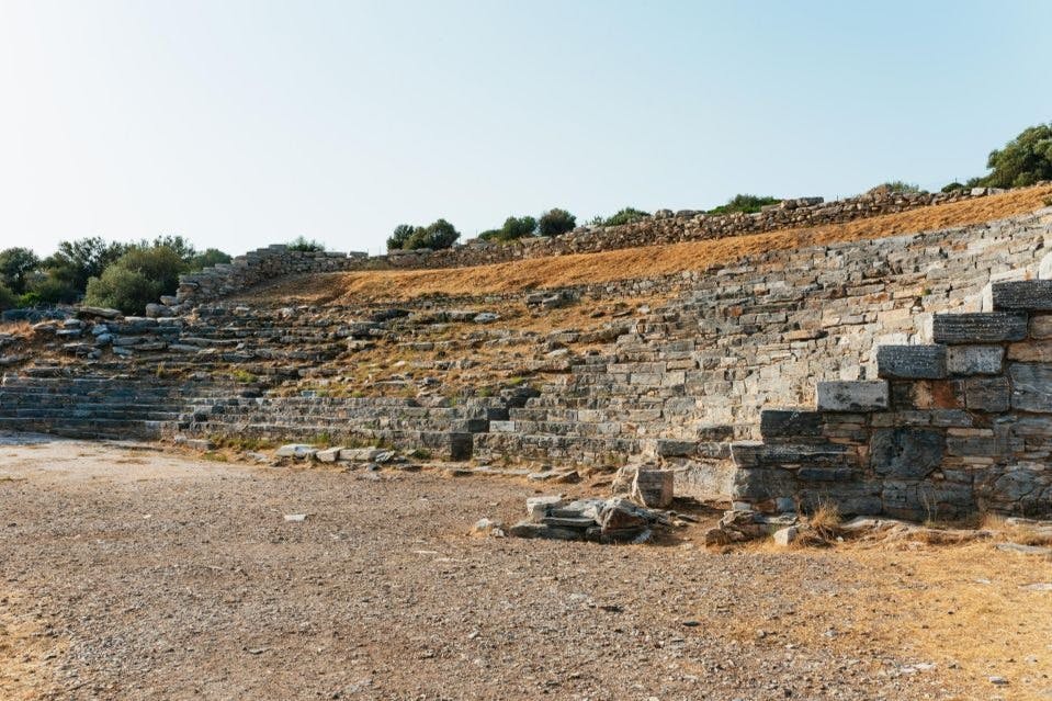 Αρχαίο πέτρινο αμφιθέατρο με σειρές από ξεθωριασμένα πέτρινα καθίσματα μια ηλιόλουστη μέρα, περιτριγυρισμένο από αραιή βλάστηση.
