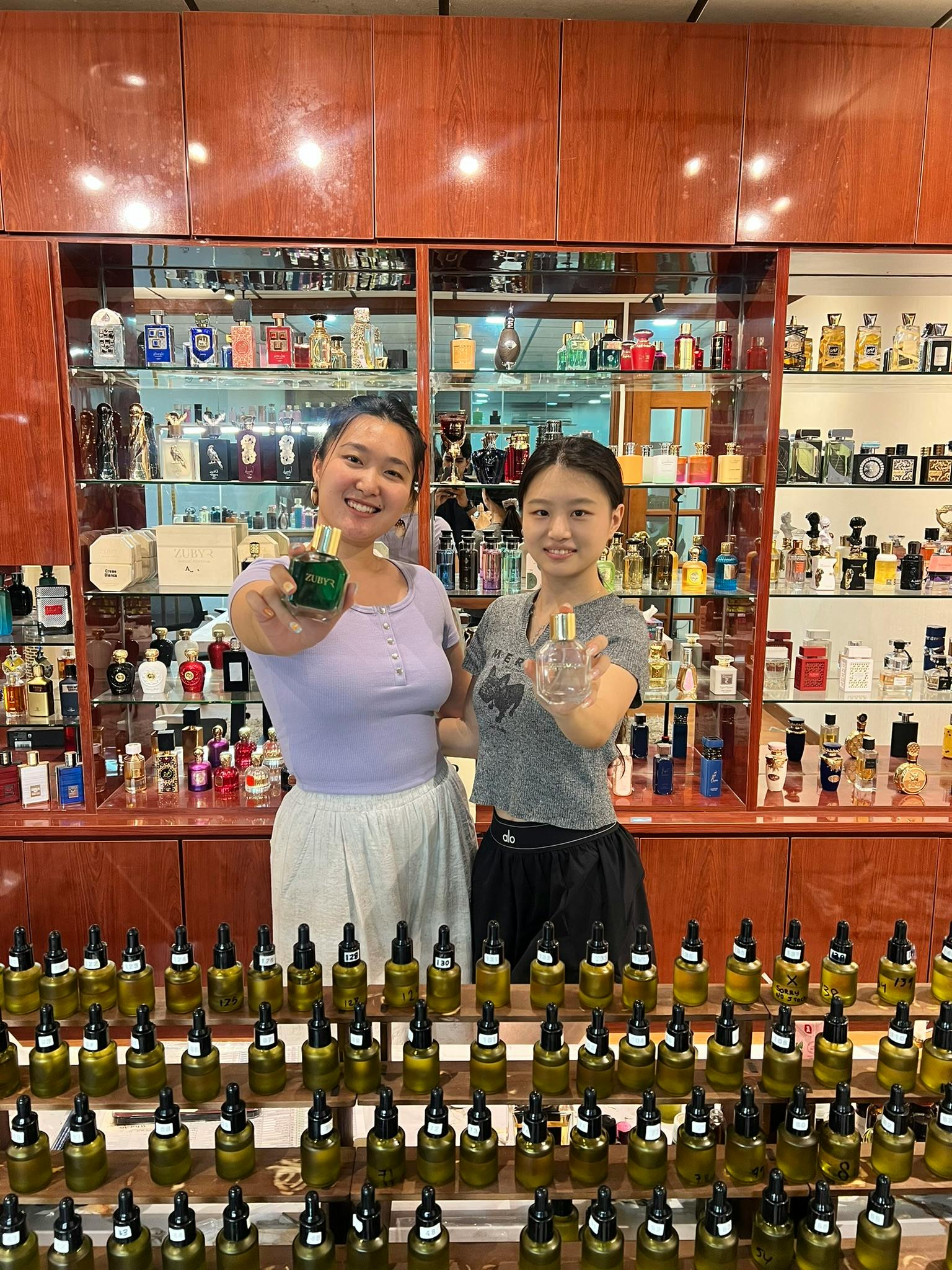 Two people holding perfume bottles in front of a display filled with various fragrances, smiling at the camera.