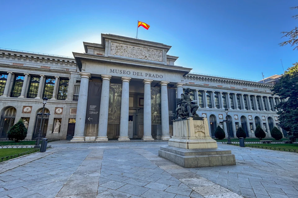 The front entrance of the Museo del Prado in Madrid with a statue in the foreground and a Spanish flag flying above.