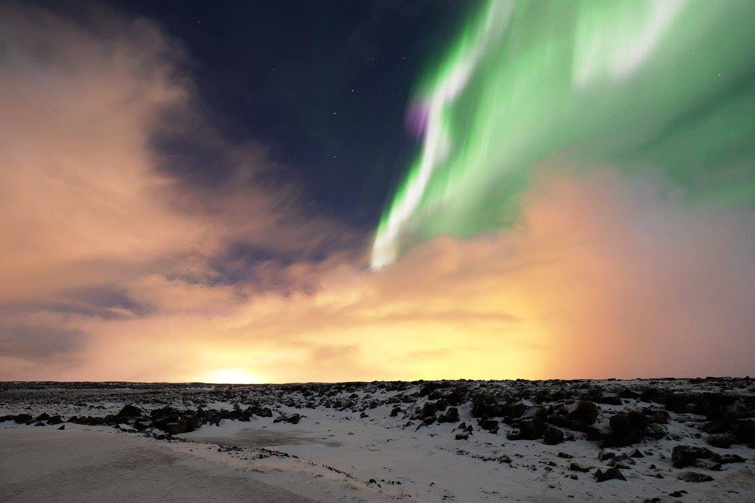 Le luci verdi dell'aurora si estendono in un cielo notturno sopra un paesaggio innevato con le nuvole illuminate da un caldo bagliore arancione.