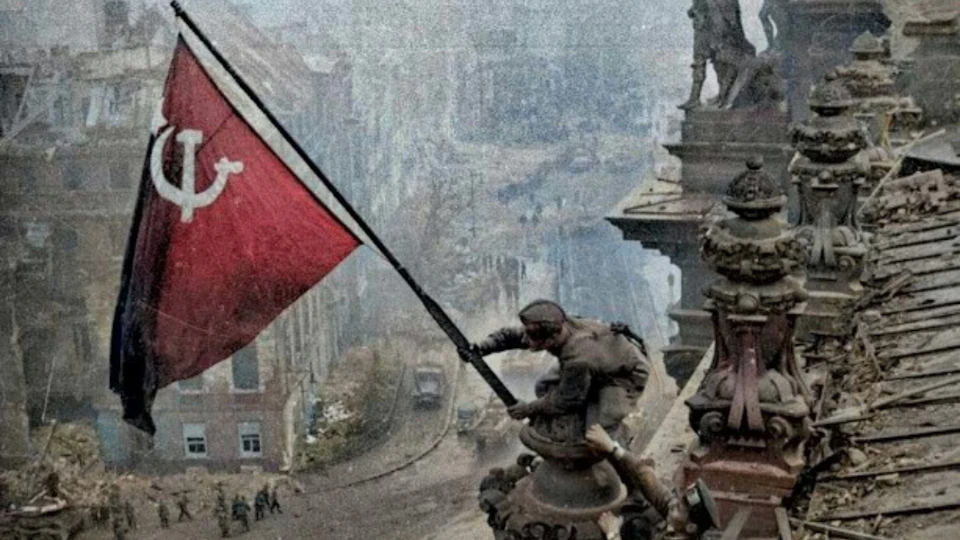 A soldier raises a red flag on a rooftop overlooking a war-torn city with rubble and damaged buildings below.