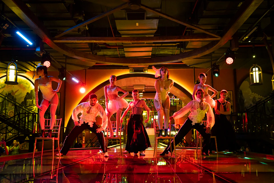 Performers in artistic costumes dancing on a lit stage with an industrial-style background and vibrant lighting.
