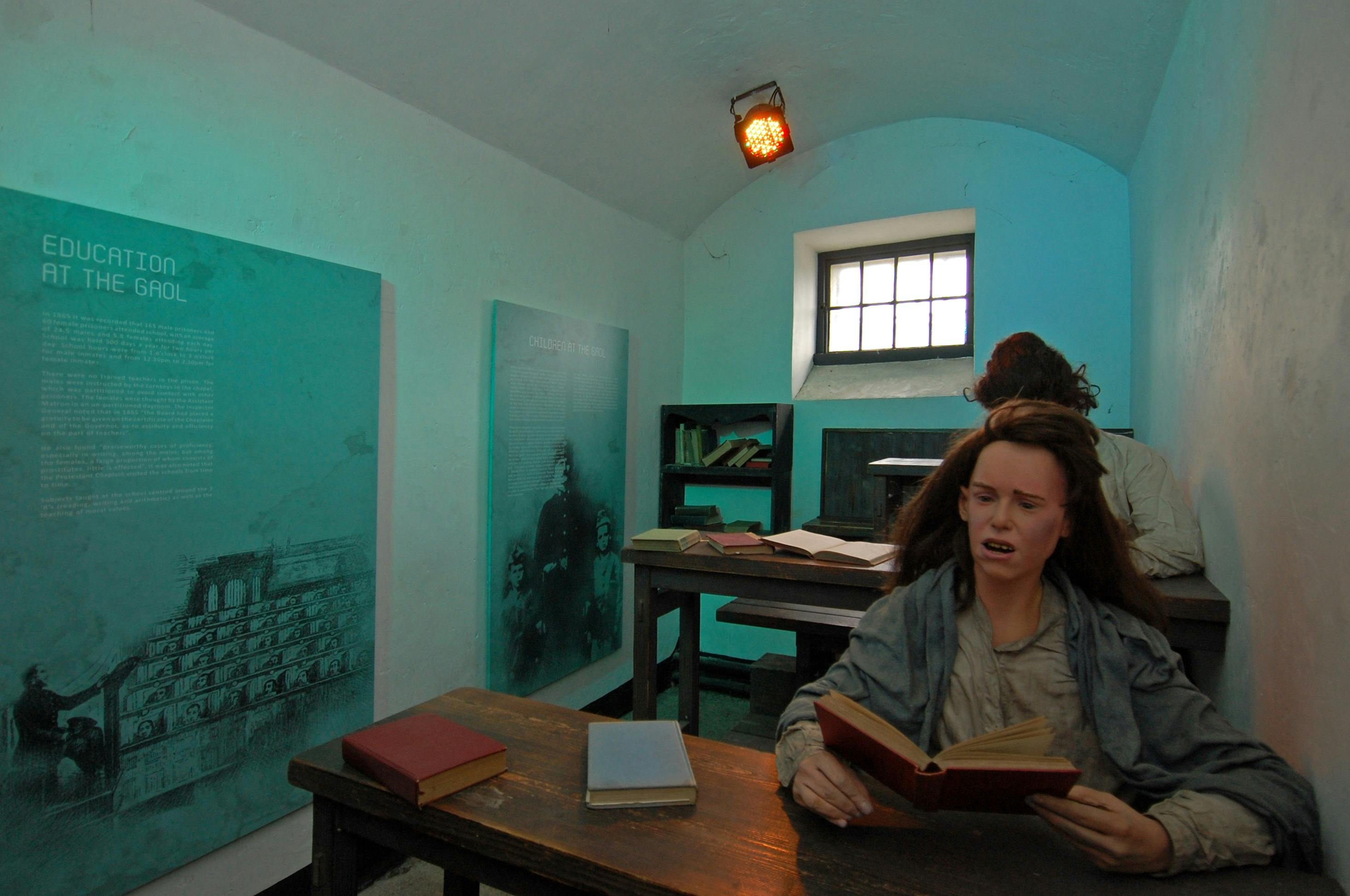 Vosková figurína sedící u stolu s otevřenou knihou v tlumeně osvětlené místnosti, která se podobá třídě nebo pracovně.