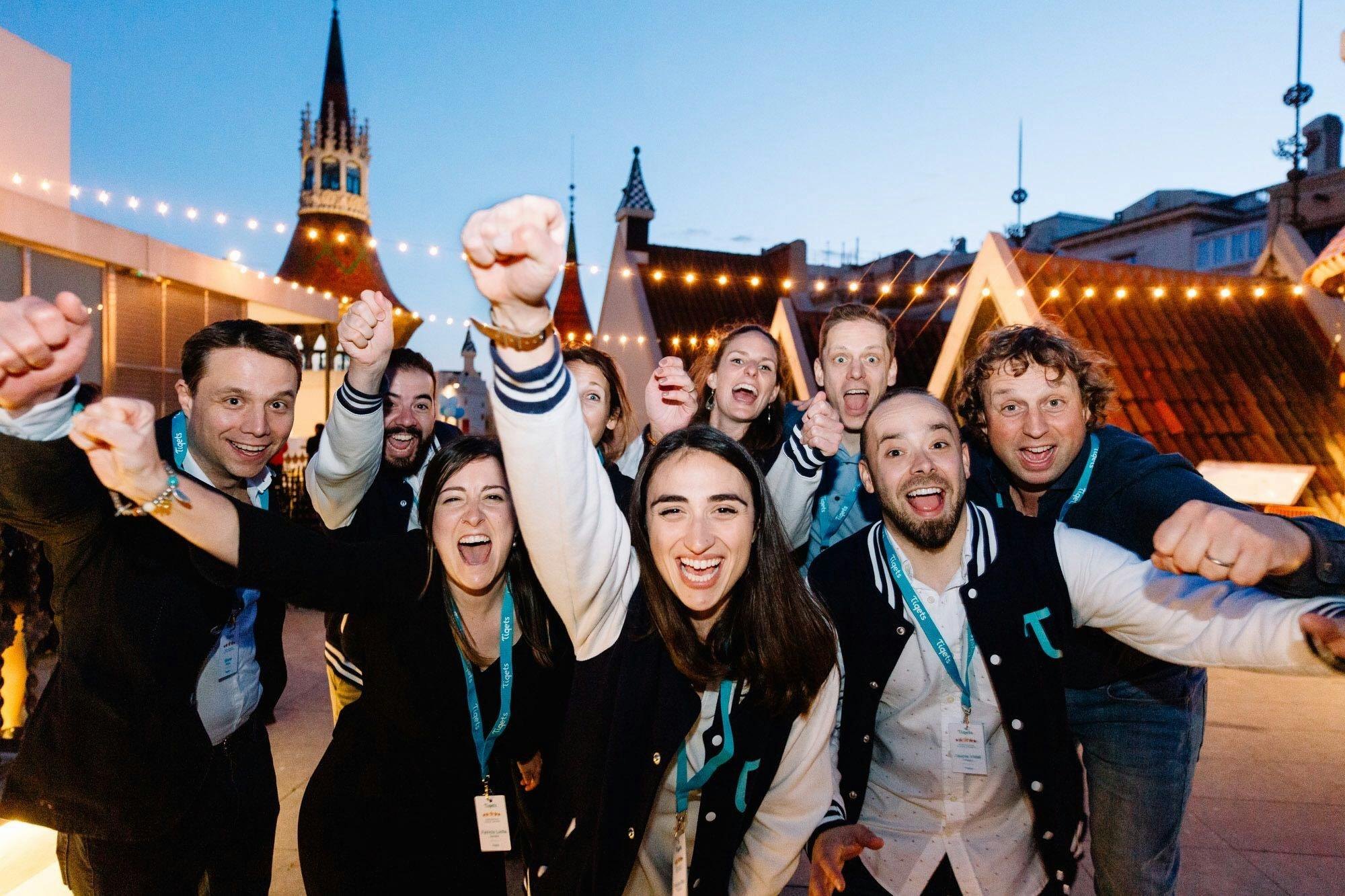 A group of people wearing matching jackets cheer enthusiastically on a rooftop with string lights and pointed towers in the background.