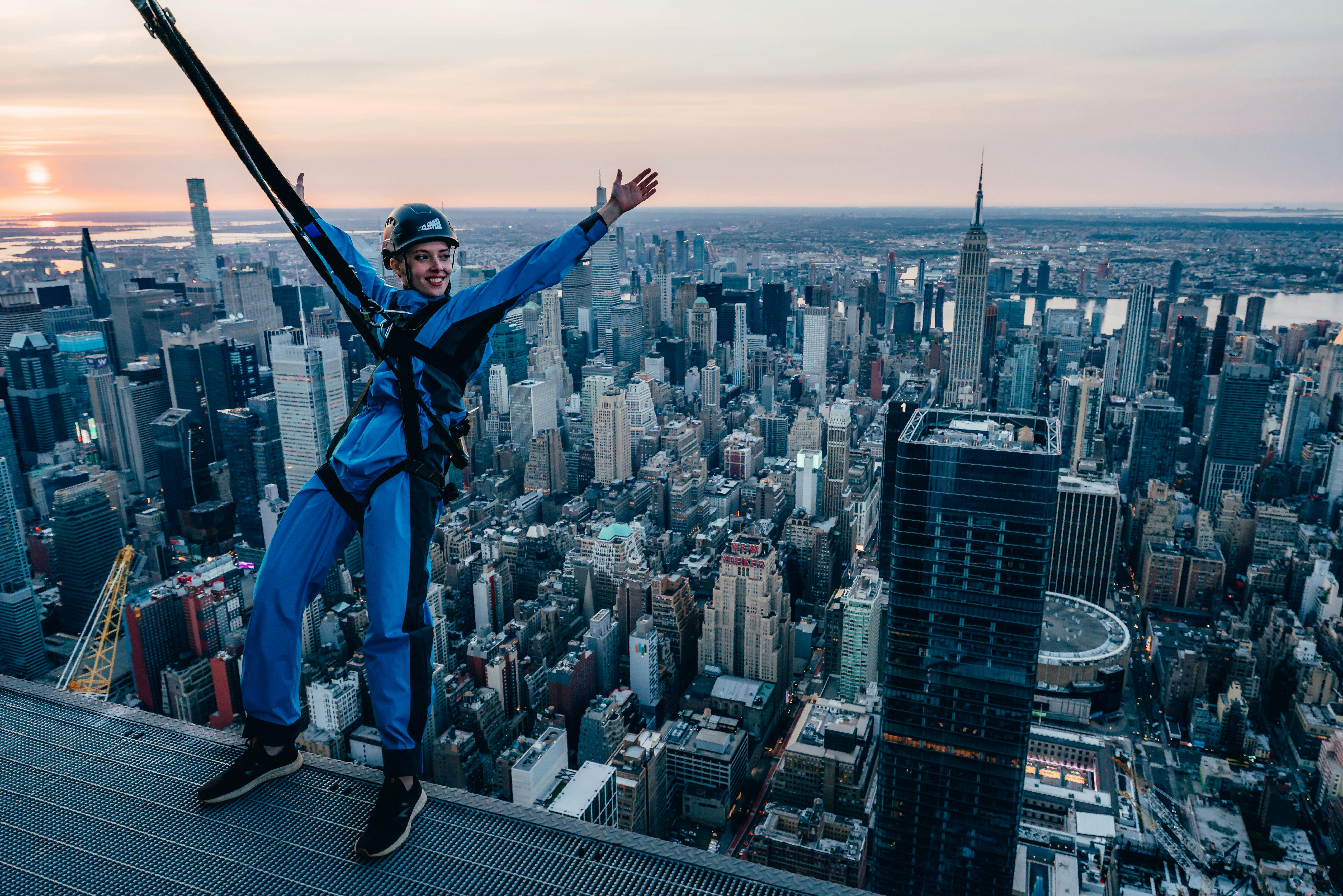 Personne vêtue d'une combinaison bleue et d'un casque, harnachée et debout sur le rebord d'un toit, les bras tendus, surplombant la ligne d'horizon d'une ville au coucher du soleil.