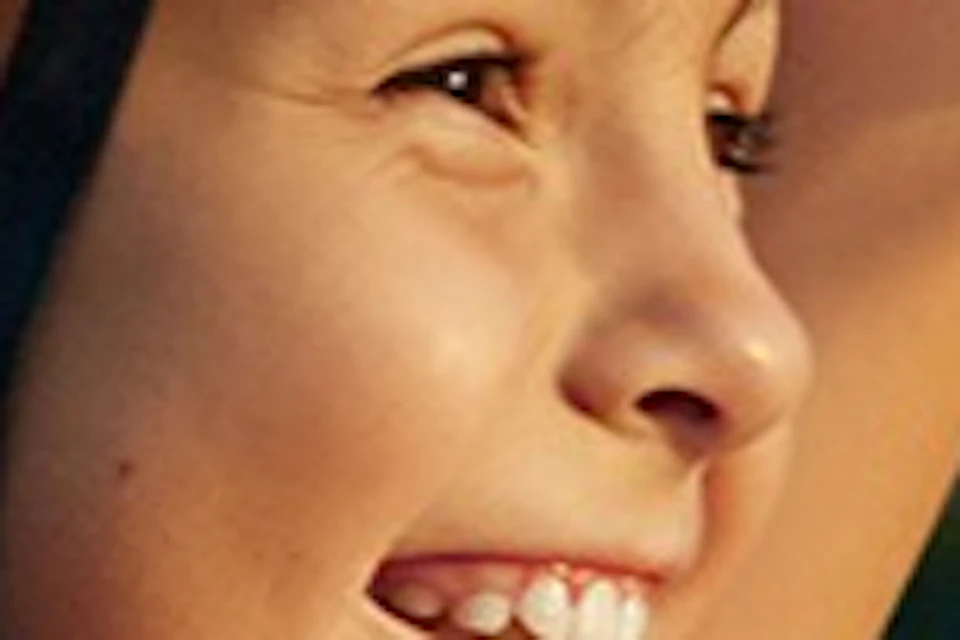 A young girl in a pink shirt and helmet smiles while zip-lining, with green trees blurred in the background.
