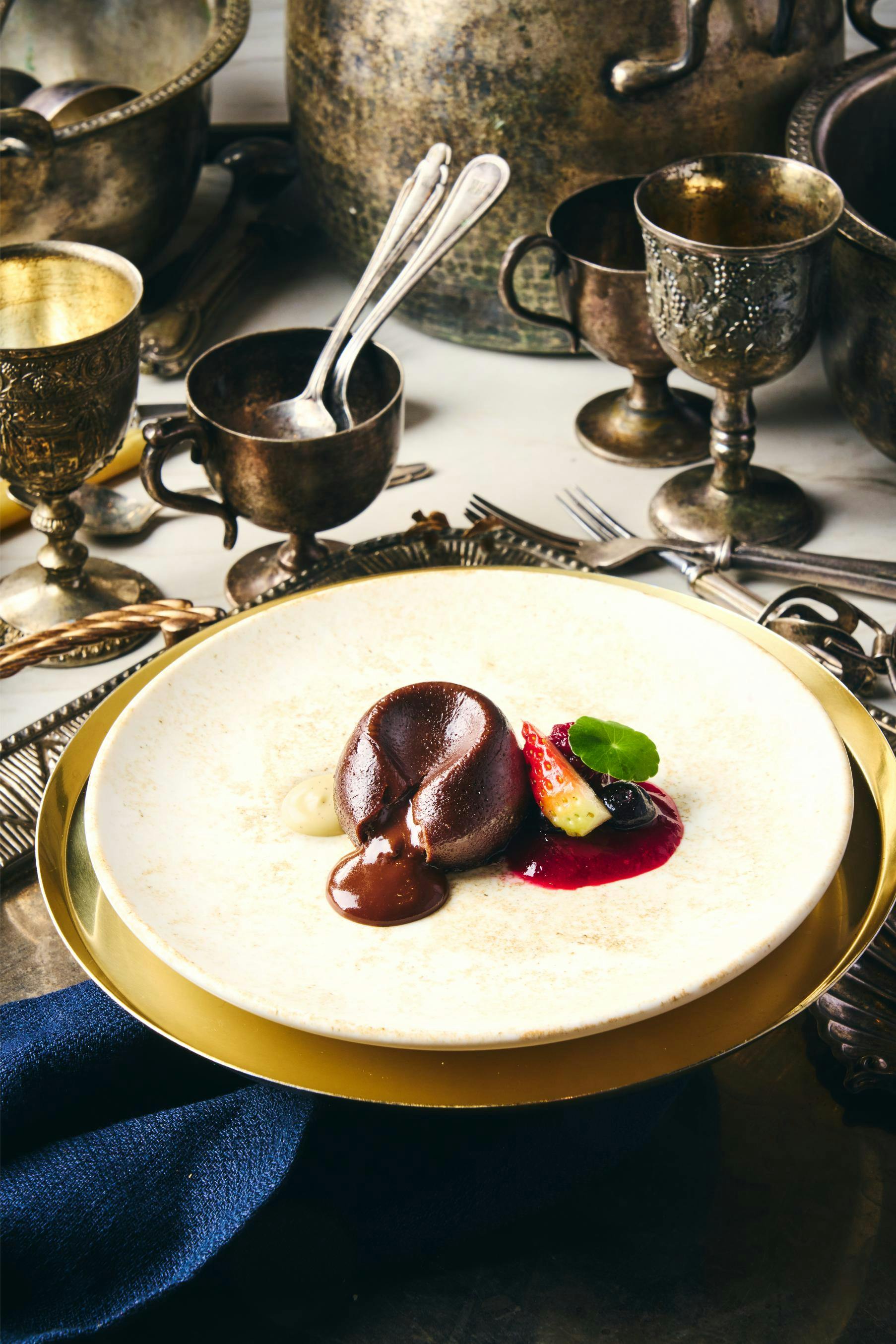 A dessert plate with chocolate mousse, berries, and sauce, surrounded by ornate cups and cutlery on a table.