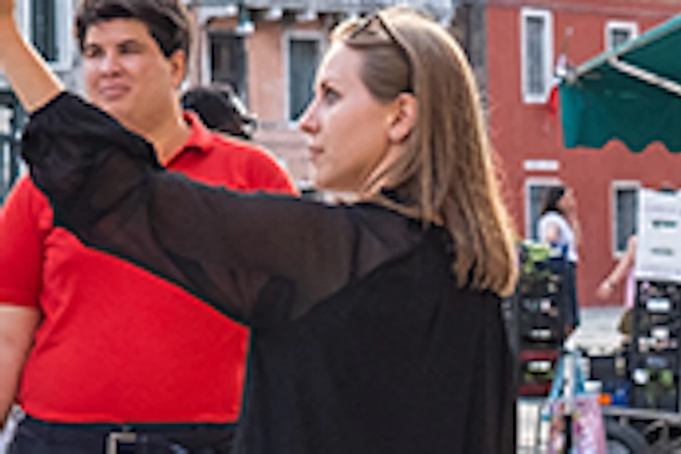 A guide wearing a black dress points as she explains something to five tourists standing in a historic square in front of old buildings.