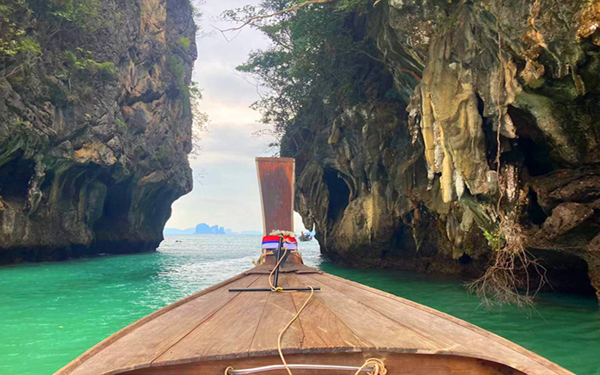 A wooden boat navigates between two tall, rocky cliffs over clear turquoise water, with distant islands visible in the background.
