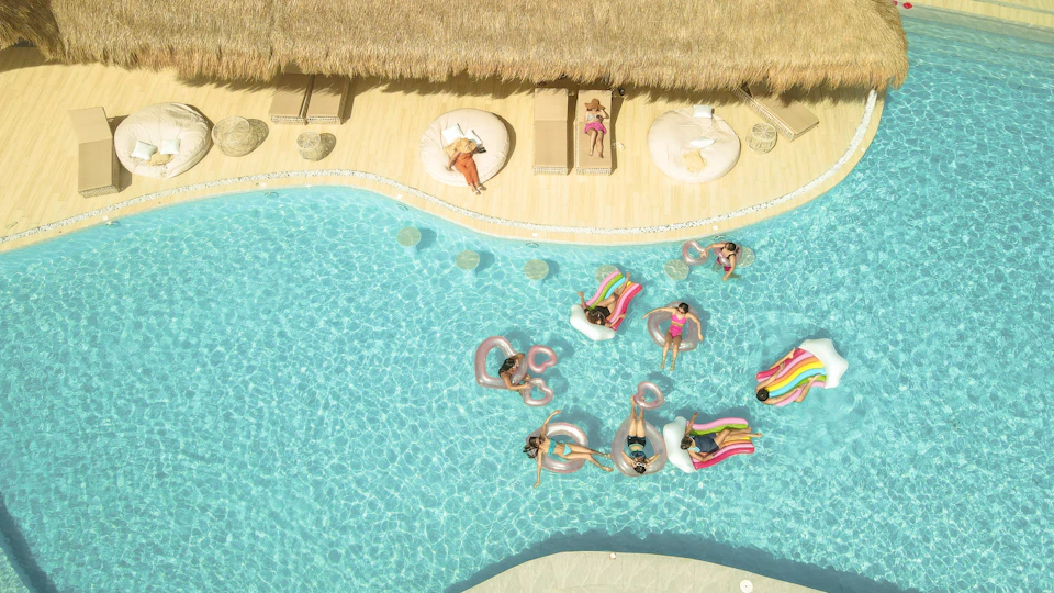 Aerial view of people relaxing on floaties in a pool, with lounge chairs and umbrellas on the deck area nearby.