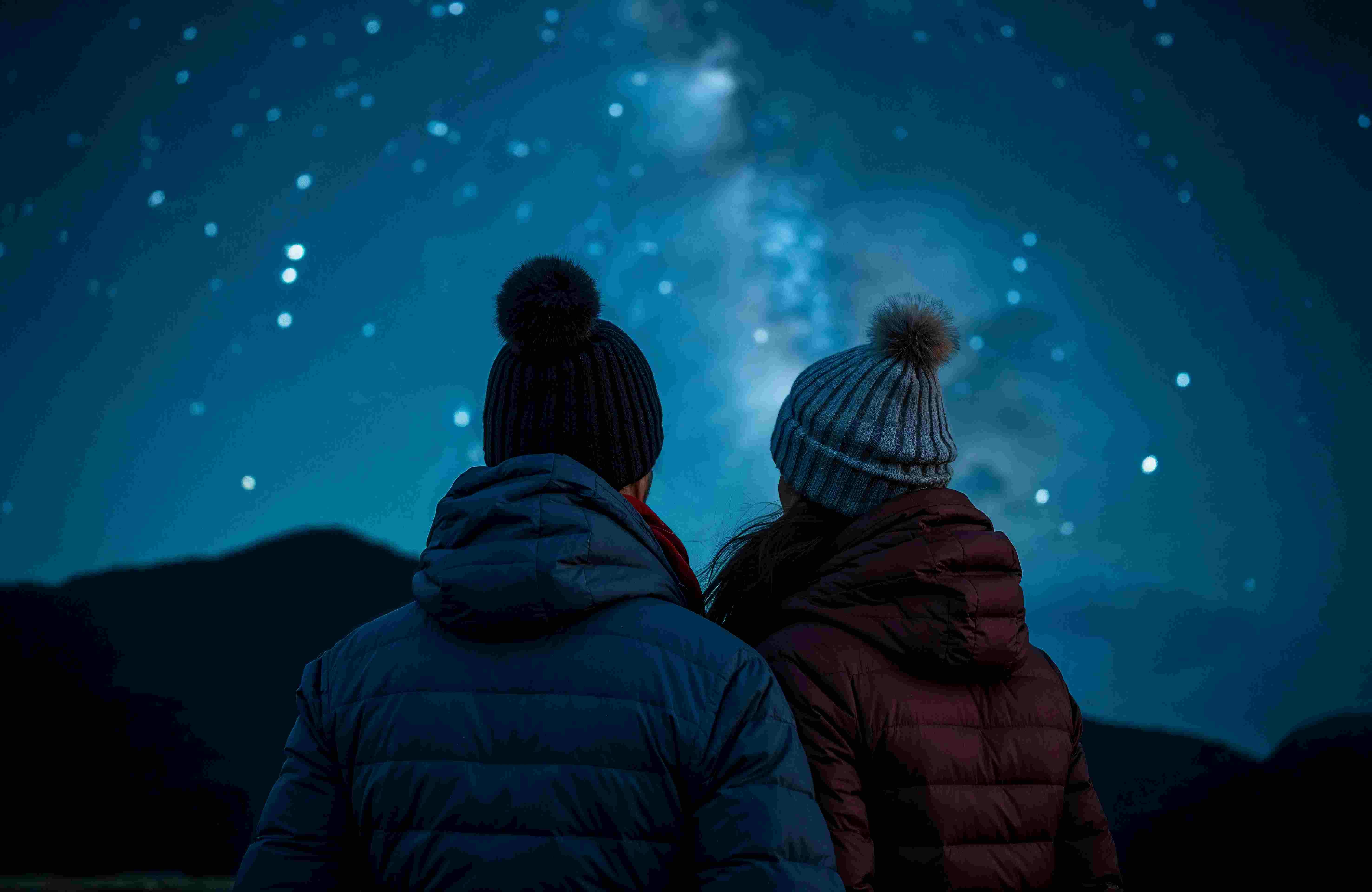 两个穿着夹克和帽子的人凝视着星光熠熠的夜空，远处是山脉的轮廓。