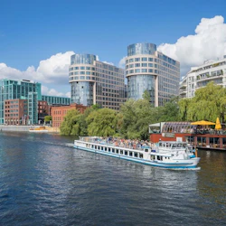 En passagerarbåt kryssar på en flod bredvid moderna byggnader och frodiga träd under en klarblå himmel med några vita moln.