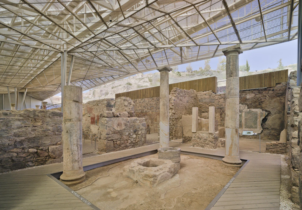 Ancient ruins with stone columns and walls under a metal and glass canopy, set in an outdoor archaeological site.