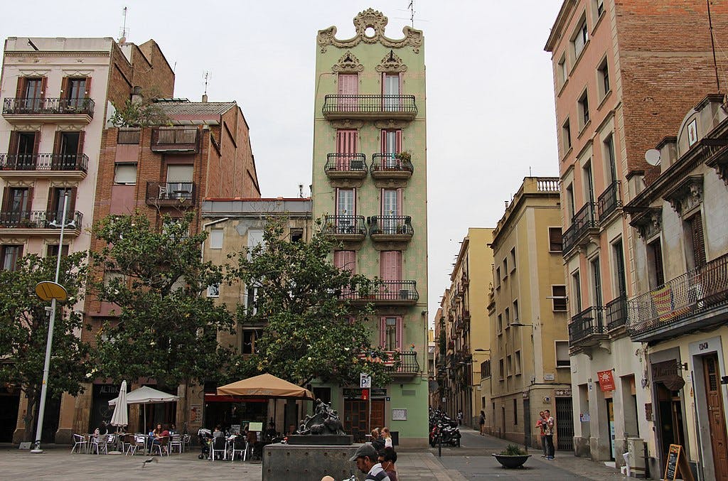 A narrow green building with intricate rooftop designs stands between taller structures in a European street, with people dining outdoors.