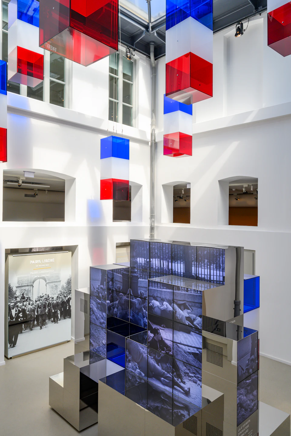 Modern art installation in a brightly lit room with suspended red, blue, and white cubes and a photo display on large screens.