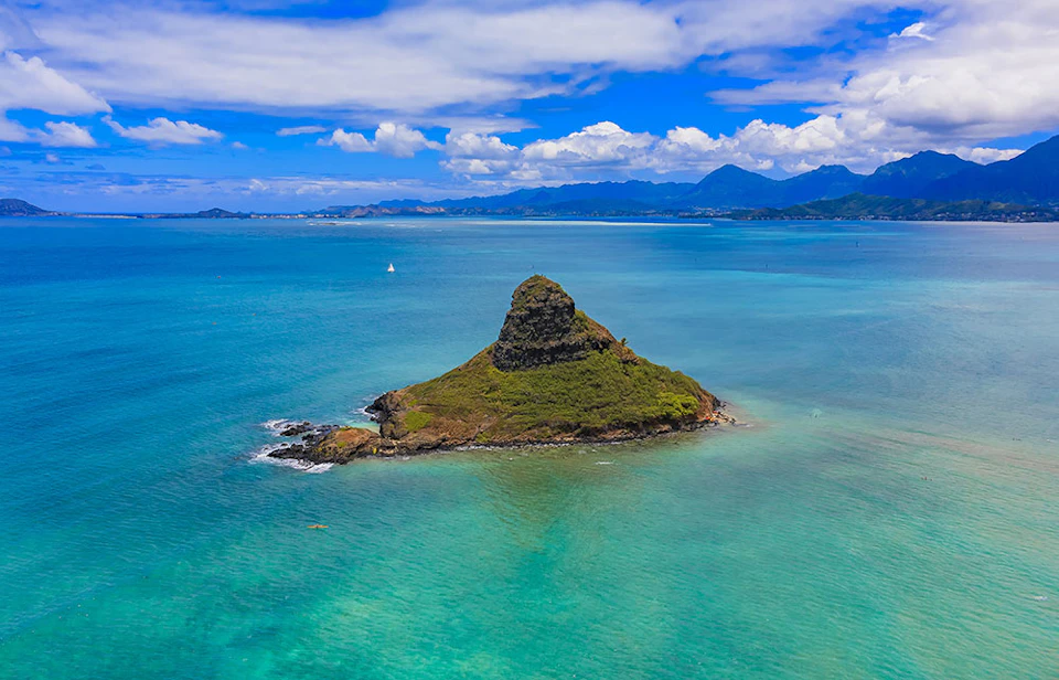 A small green island with a conical peak surrounded by turquoise waters under a partly cloudy sky, with distant mountains in the background.
