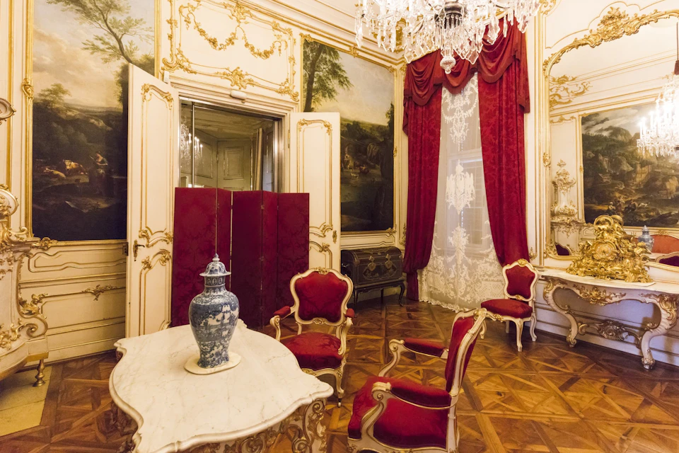 Ornate room with red velvet chairs, marble table with a blue vase, chandelier, and large red drapes over lace curtains.