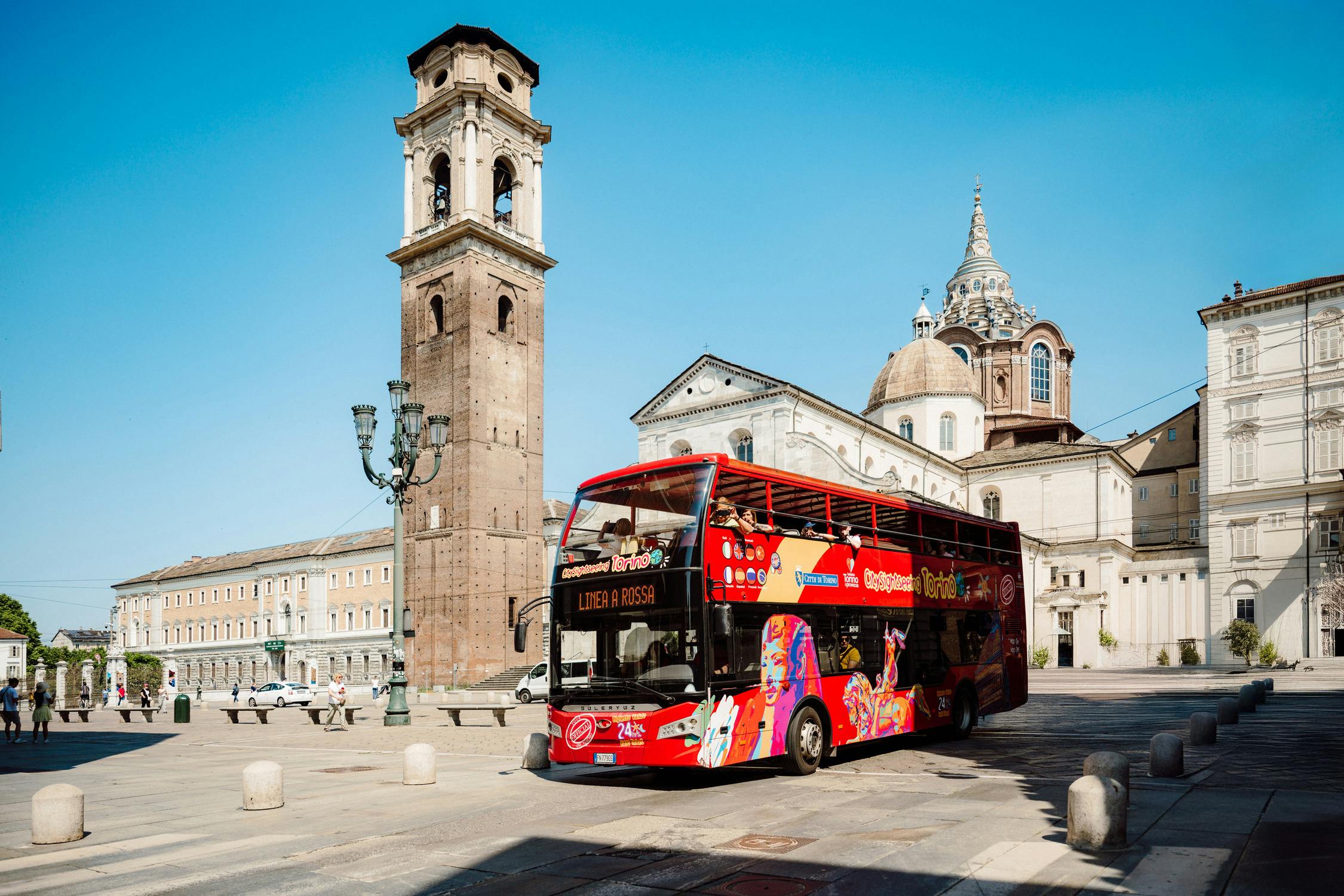 A red double-decker tour bus near a historic church and bell tower in a sunny plaza.