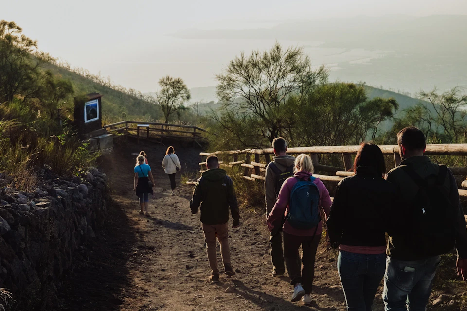 A group of people walking down a dirt path on a hillside, bordered by wooden fences and sparse trees.