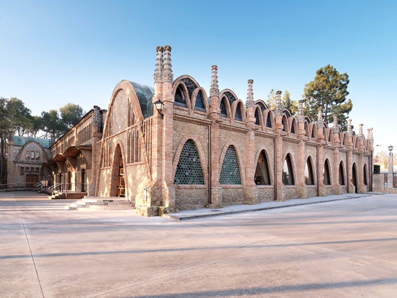 Un long bâtiment historique avec de multiples fenêtres cintrées et des cheminées, entouré d'arbres sous un ciel bleu clair.