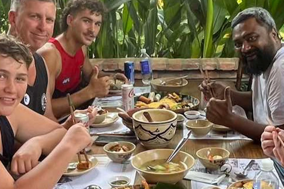Six people seated at a wooden table enjoying a meal outdoors, surrounded by lush greenery and brick columns.