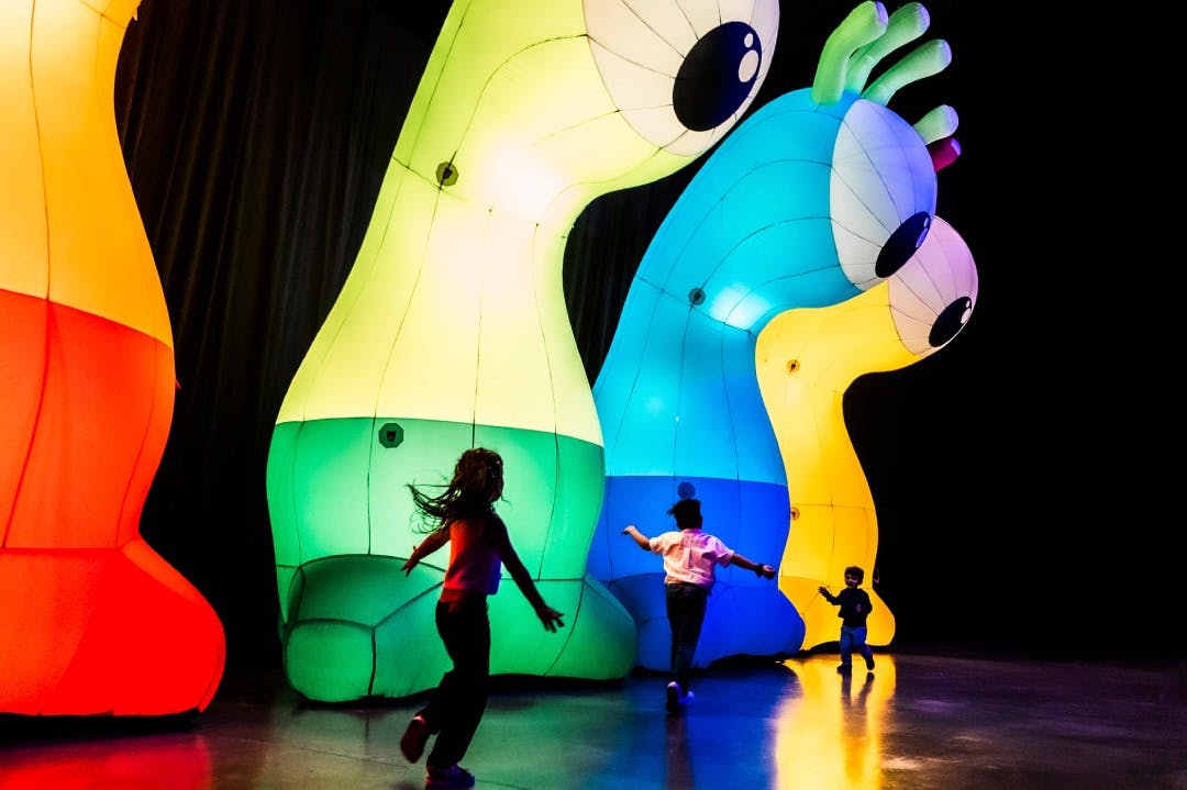 Des enfants courent près de grandes structures gonflables colorées et illuminées, qui ressemblent à des créatures abstraites, dans une pièce sombre.