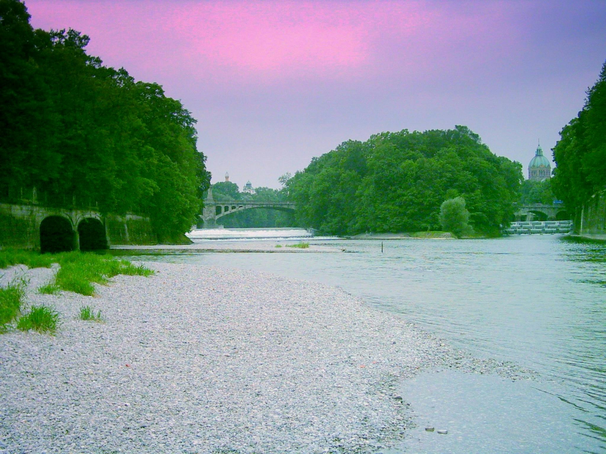 Un fiume tranquillo con una riva di ciottoli, alberi verdi e rigogliosi, ponti e un edificio a cupola in lontananza sotto un cielo rosa.