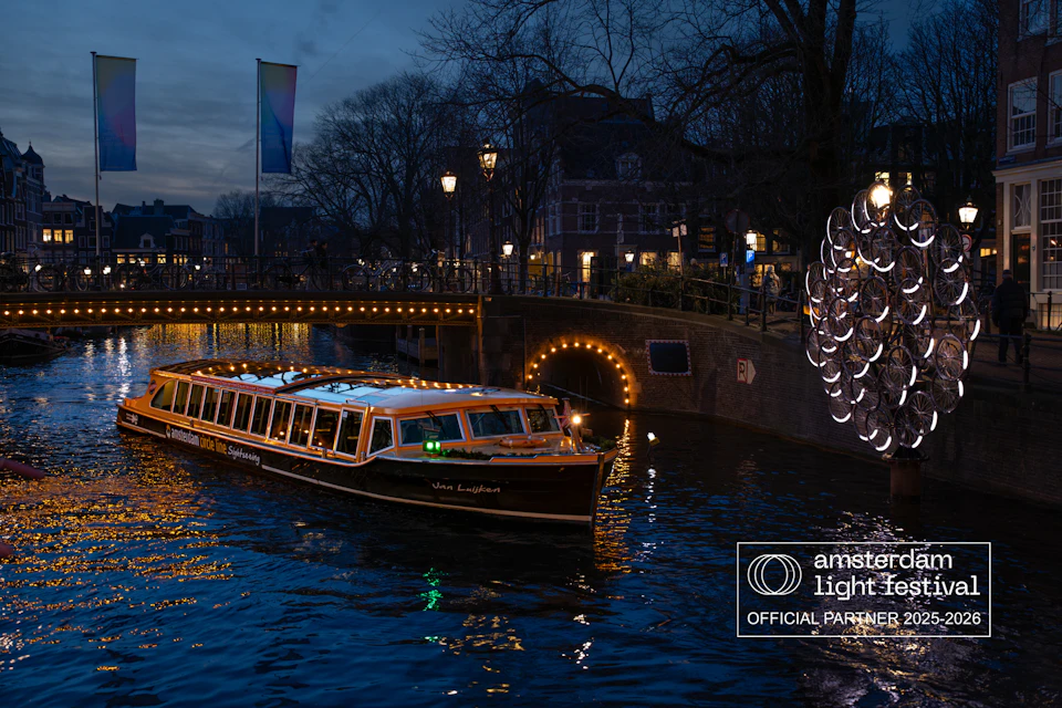 A boat decorated with lights travels on a canal at night, with a bridge and illuminated sculpture in the background.