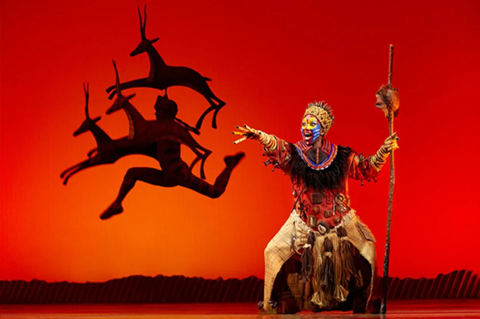 A theatrical performance featuring an actor in elaborate costume and makeup gesturing toward a silhouette of antelopes on stage.