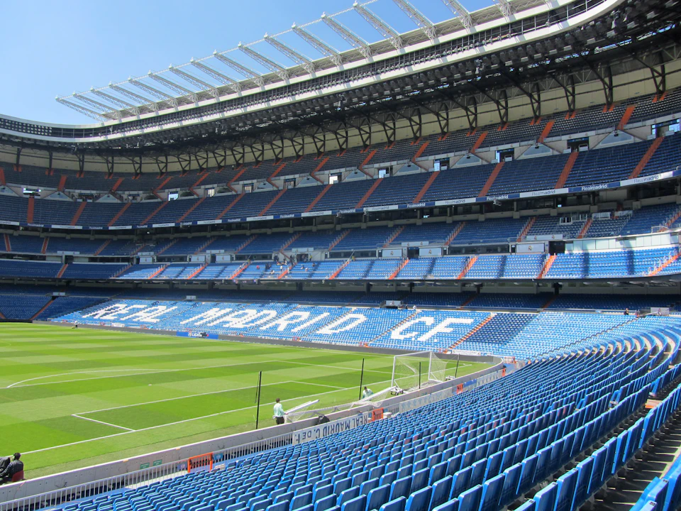 Empty football stadium with blue seats and "Real Madrid C.F." lettering. Green field and clear blue sky.