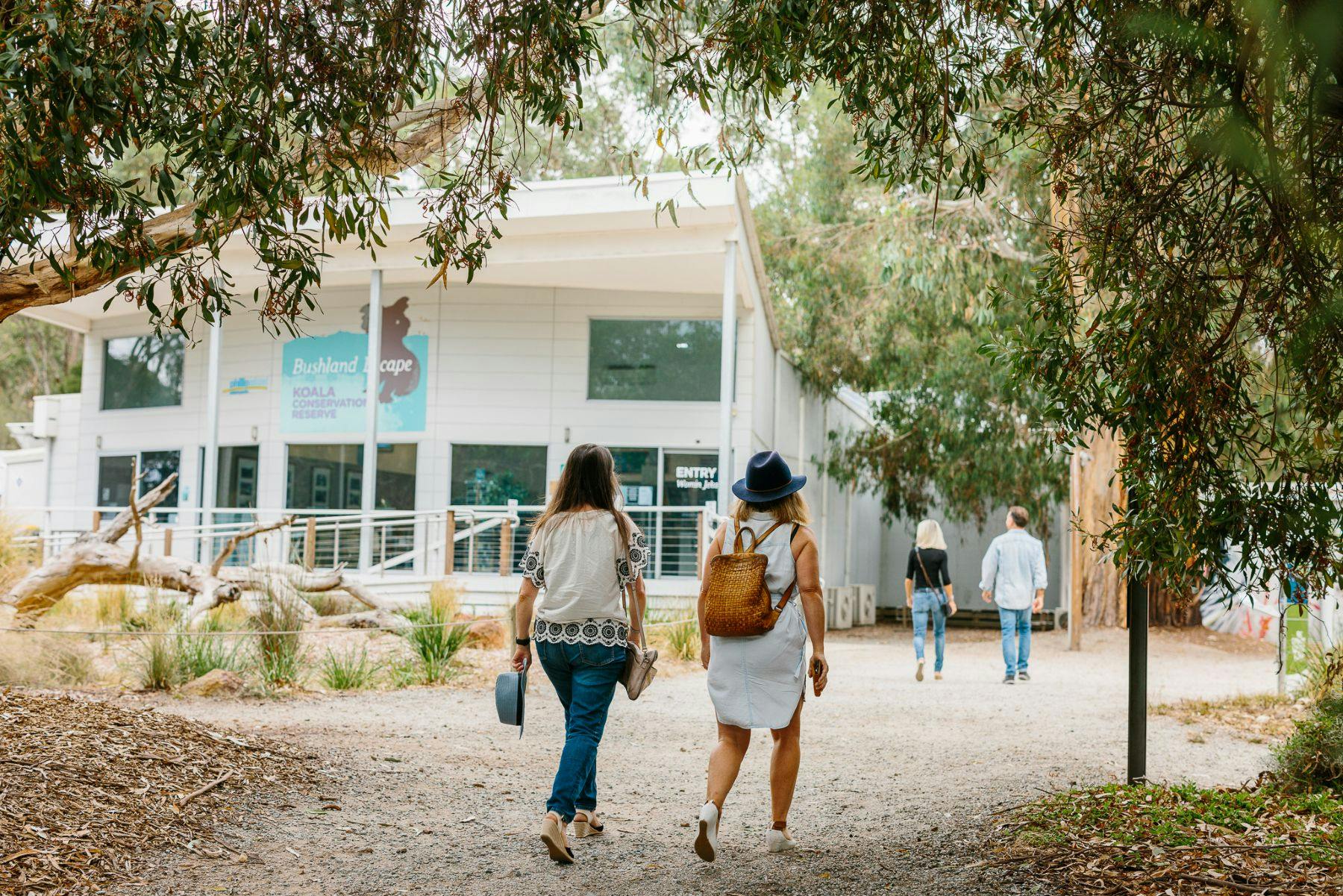 Two women walking towards a building surrounded by trees; one carries a backpack, the other holds a bag and hat. Two other people in front.
