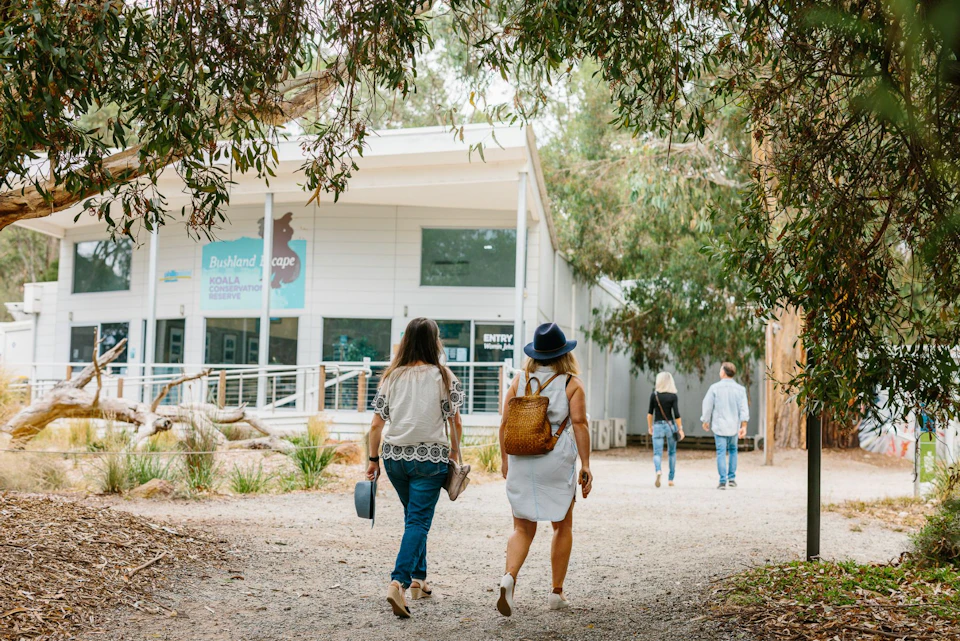 Two women walking towards a building surrounded by trees; one carries a backpack, the other holds a bag and hat. Two other people in front.