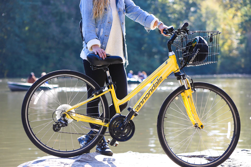 Person standing with a yellow Cannondale bicycle near a body of water, dressed in black pants, light sweater, and a denim jacket.