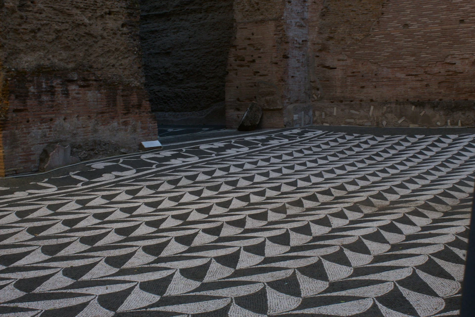 A black-and-white geometric mosaic floor pattern next to rough brown brick walls and a small sign on the ground.