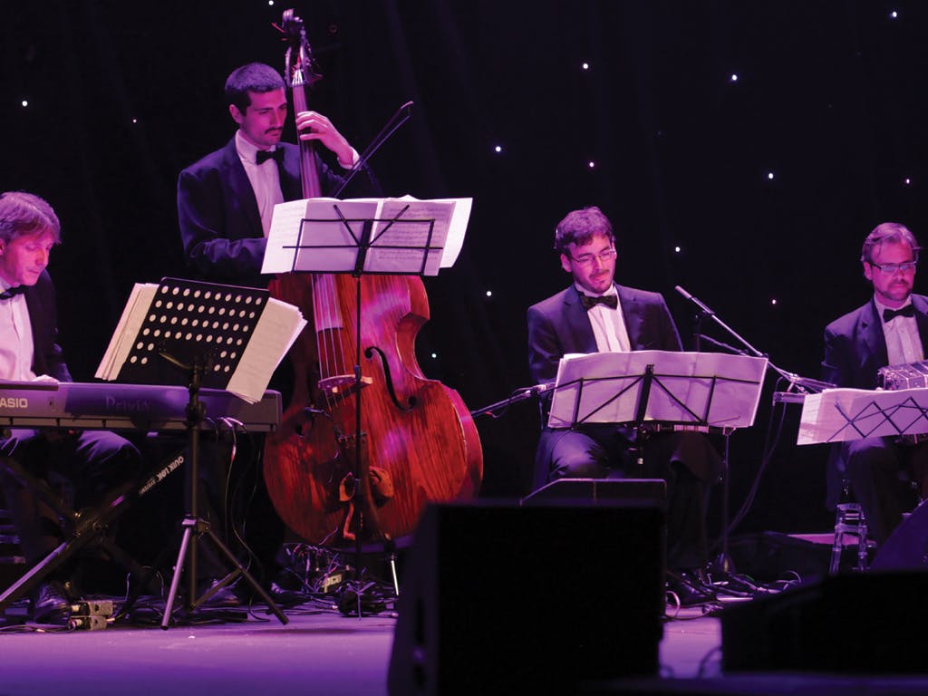 Three musicians in tuxedos perform on stage with a keyboard, double bass, and sheet music stands in front of a black backdrop with lights.