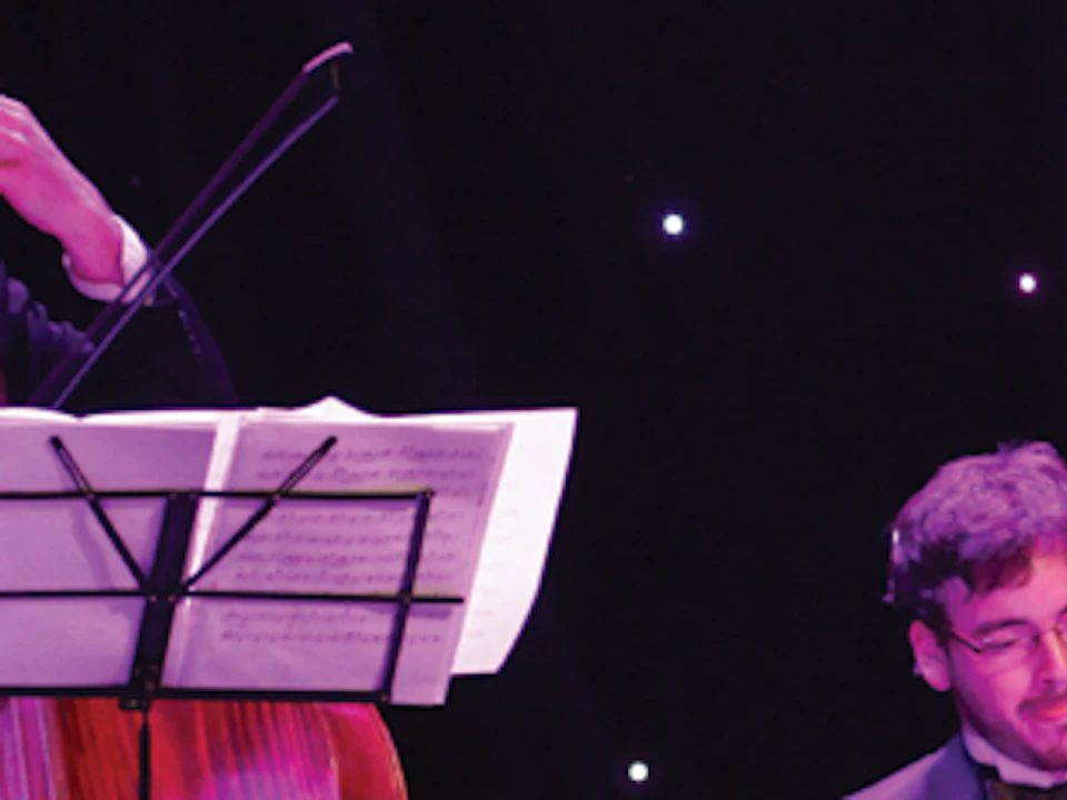 Three musicians in tuxedos perform on stage with a keyboard, double bass, and sheet music stands in front of a black backdrop with lights.
