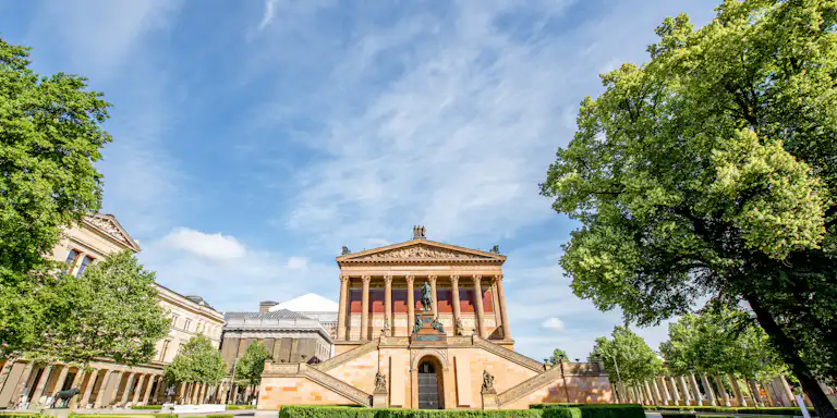 Ornate historic building with columns and stairs, surrounded by manicured lawns and trees under a partly cloudy blue sky.