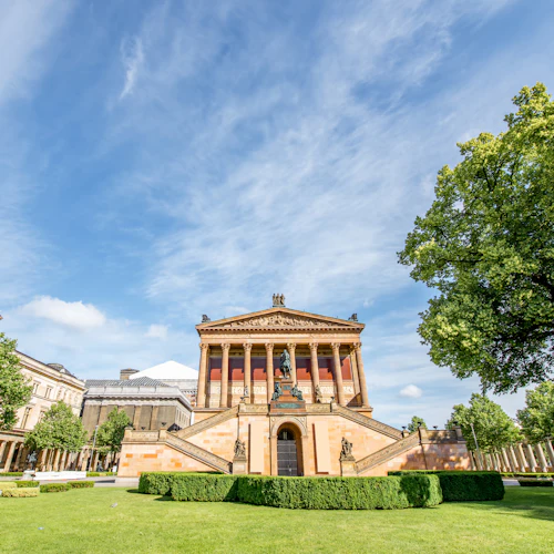 Utsmyckad historisk byggnad med pelare och trappor, omgiven av välskötta gräsmattor och träd under en delvis molnig blå himmel.