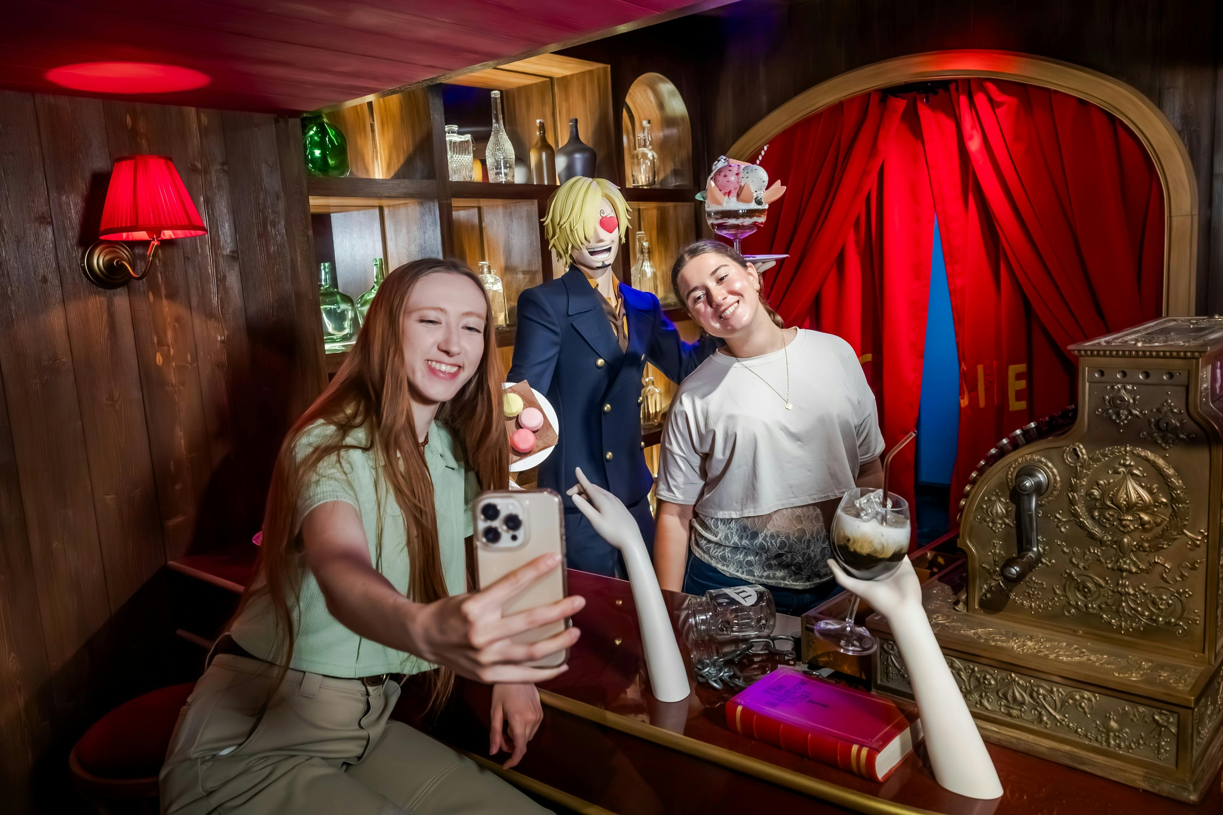 Två kvinnor tar en selfie på en träbar, med en skyltdocka av blond karaktär och olika föremål på hyllorna i bakgrunden.