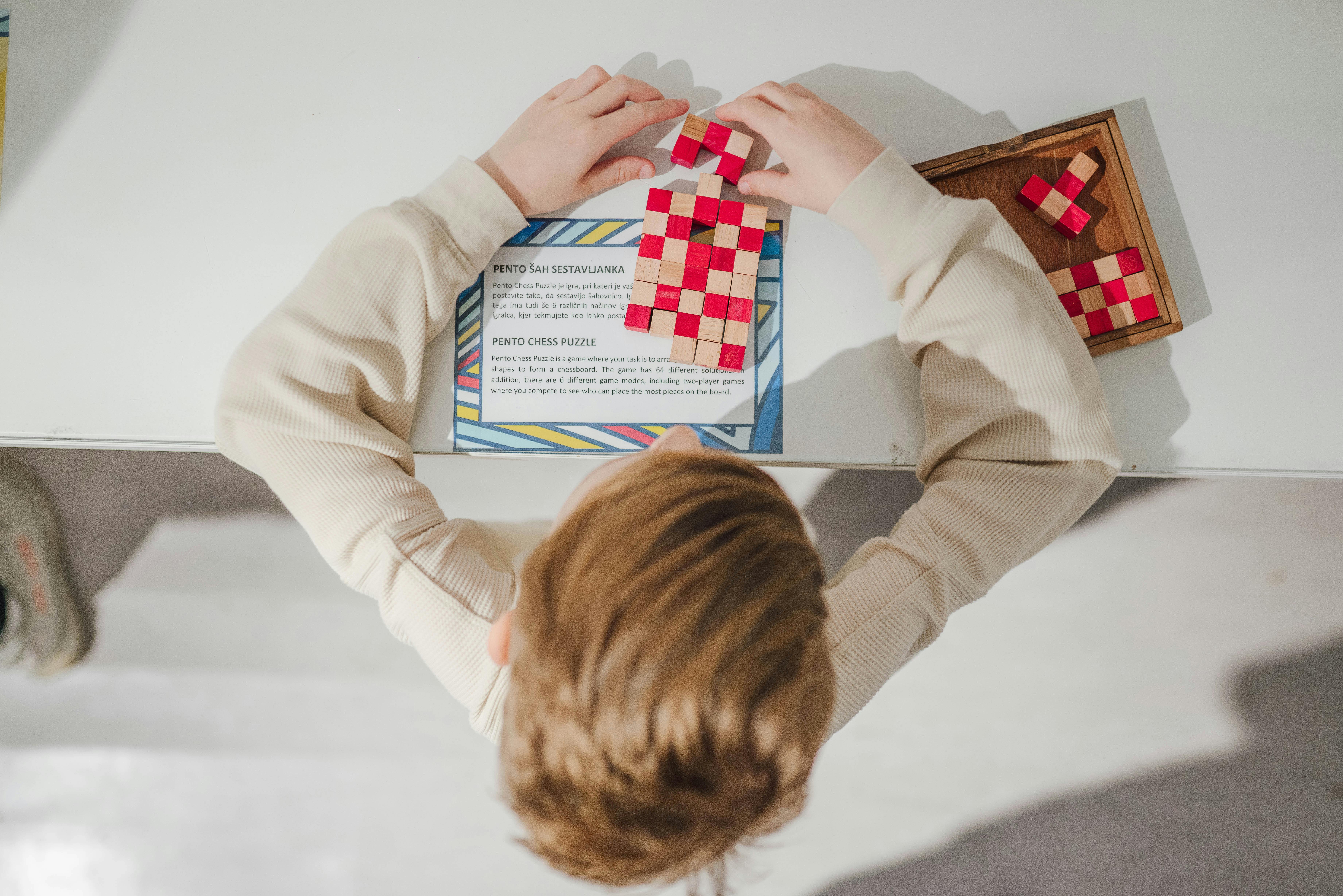 Enfant assemblant un puzzle à carreaux rouges et blancs sur une table, avec un livret ouvert devant lui et d'autres pièces dans un plateau en bois.