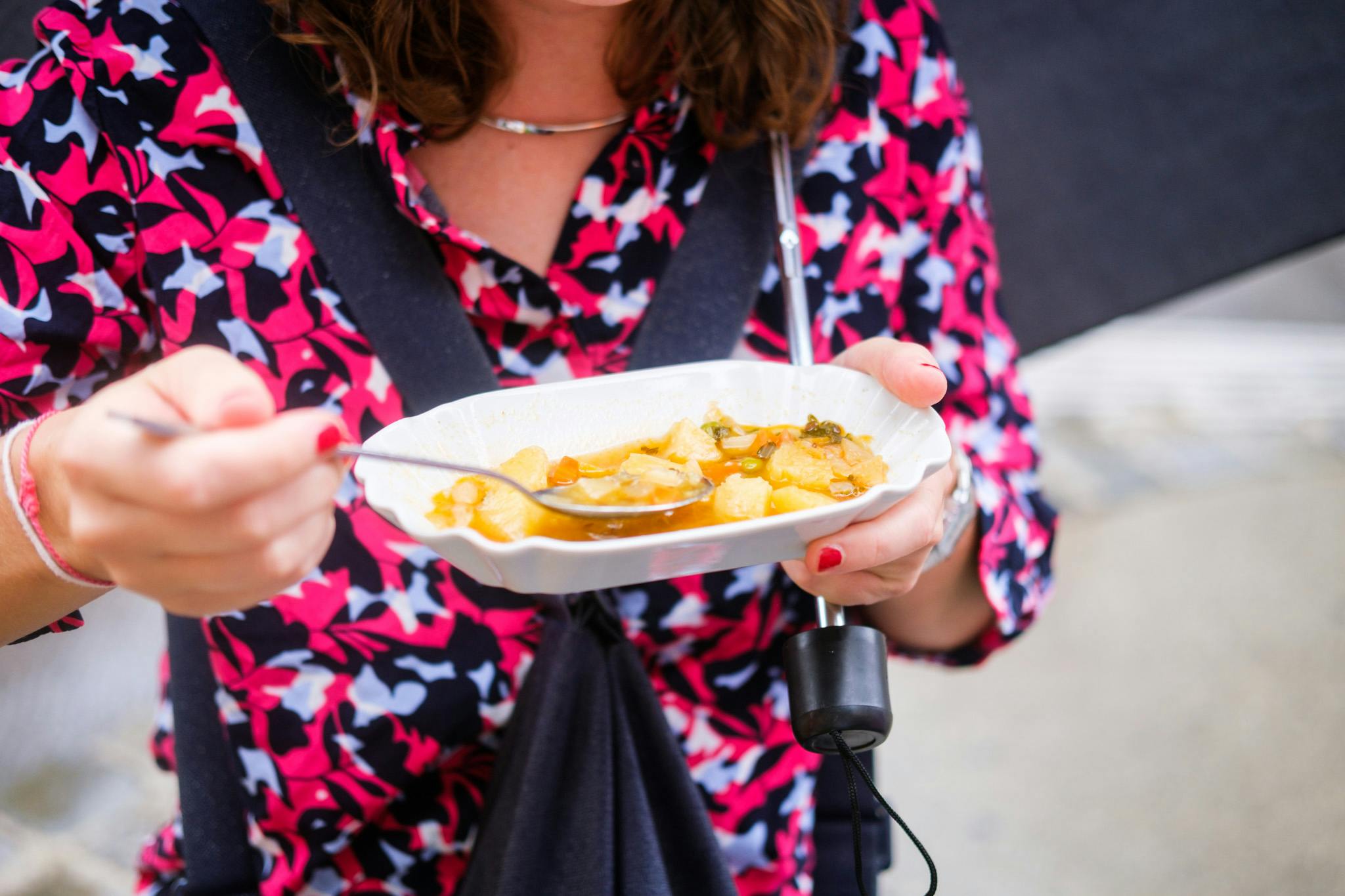 Osoba v růžovo-černé vzorované košili drží deštník a bílý talíř s jídlem a jí lžící.