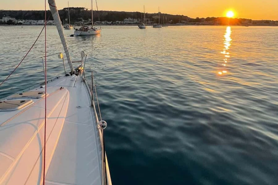 View from a sailboat's deck overlooking calm water with several other boats in the distance and the sun setting on the horizon.