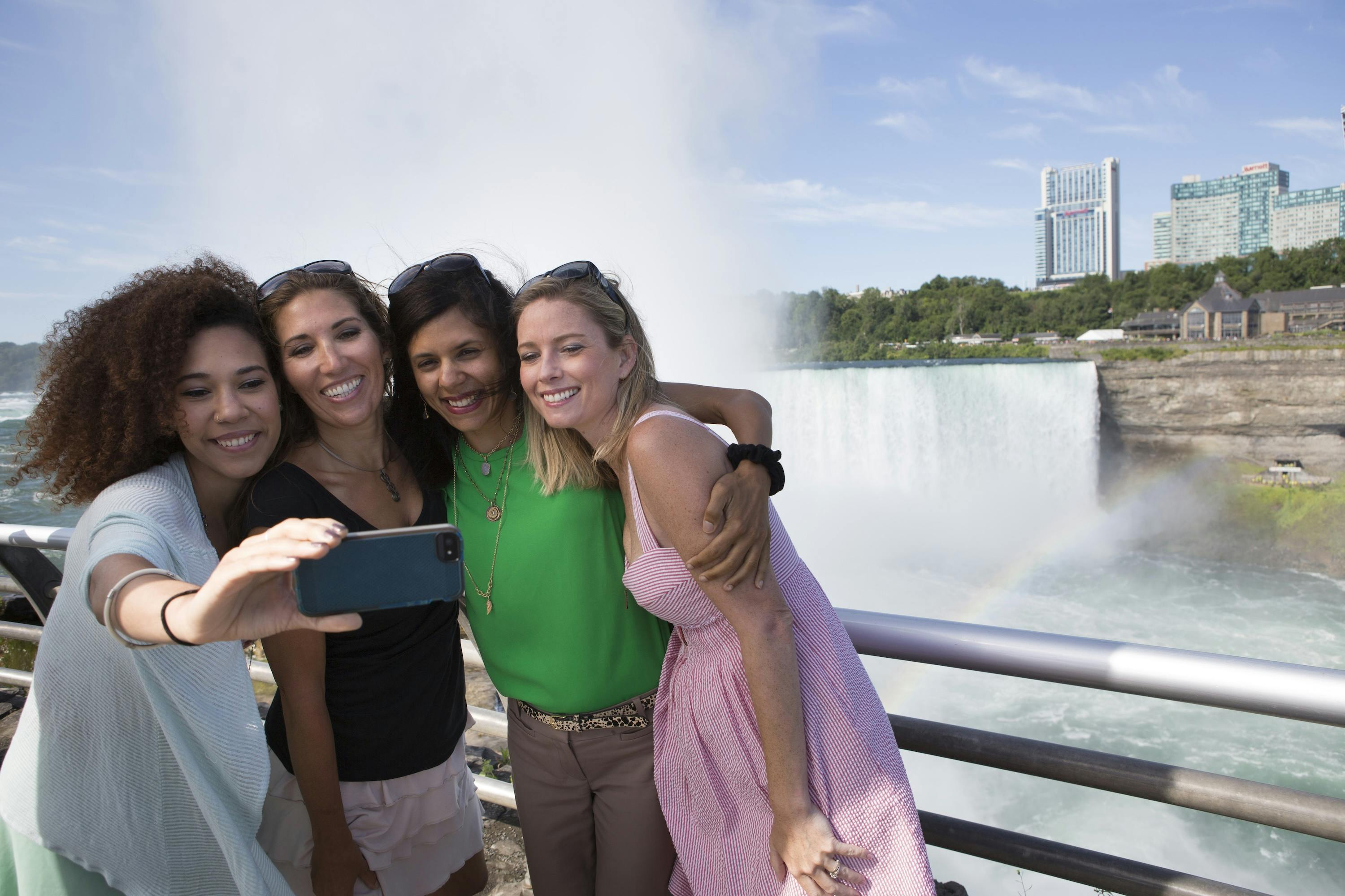 四位女性在瀑布前自拍，面带微笑。 天空晴朗，有一些白云。